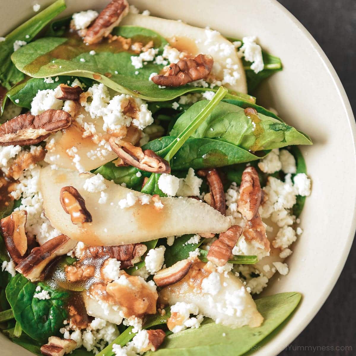 A beige bowl with baby spinach, slices of pear, crumbled goat cheese and pecans.