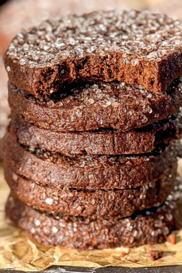 A stack of 6 chocolate shortbread cookies on parchment paper. The top cookie has a bite taken out of it. Photo by Cynthia from But First We Brunch.
