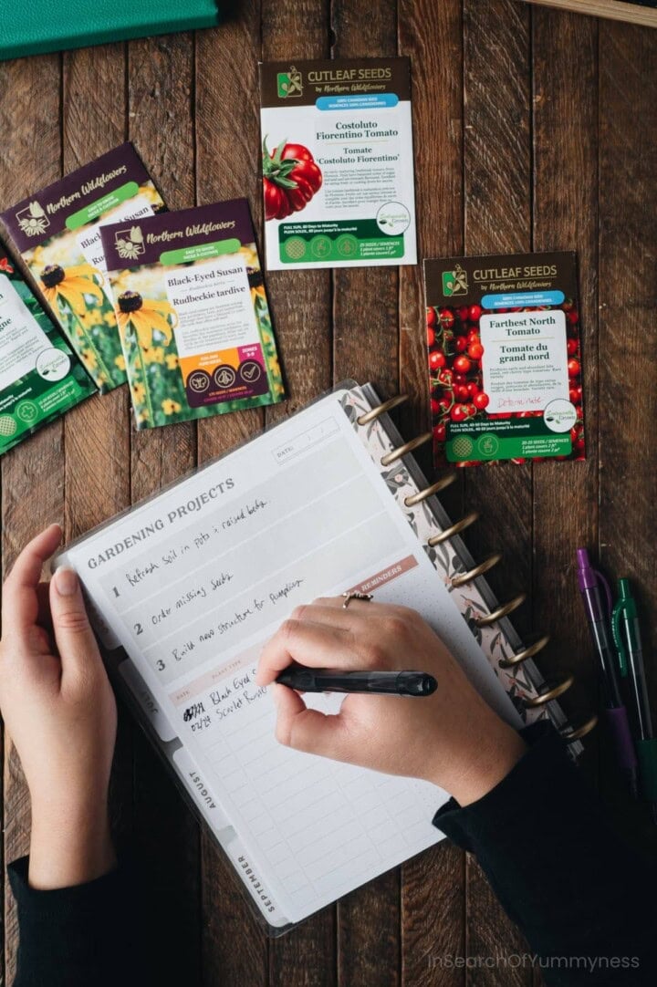 A hand writing in a gardening projects workbook next to packets of Northern Wildflowers and Cutleaf Seeds garden seeds.