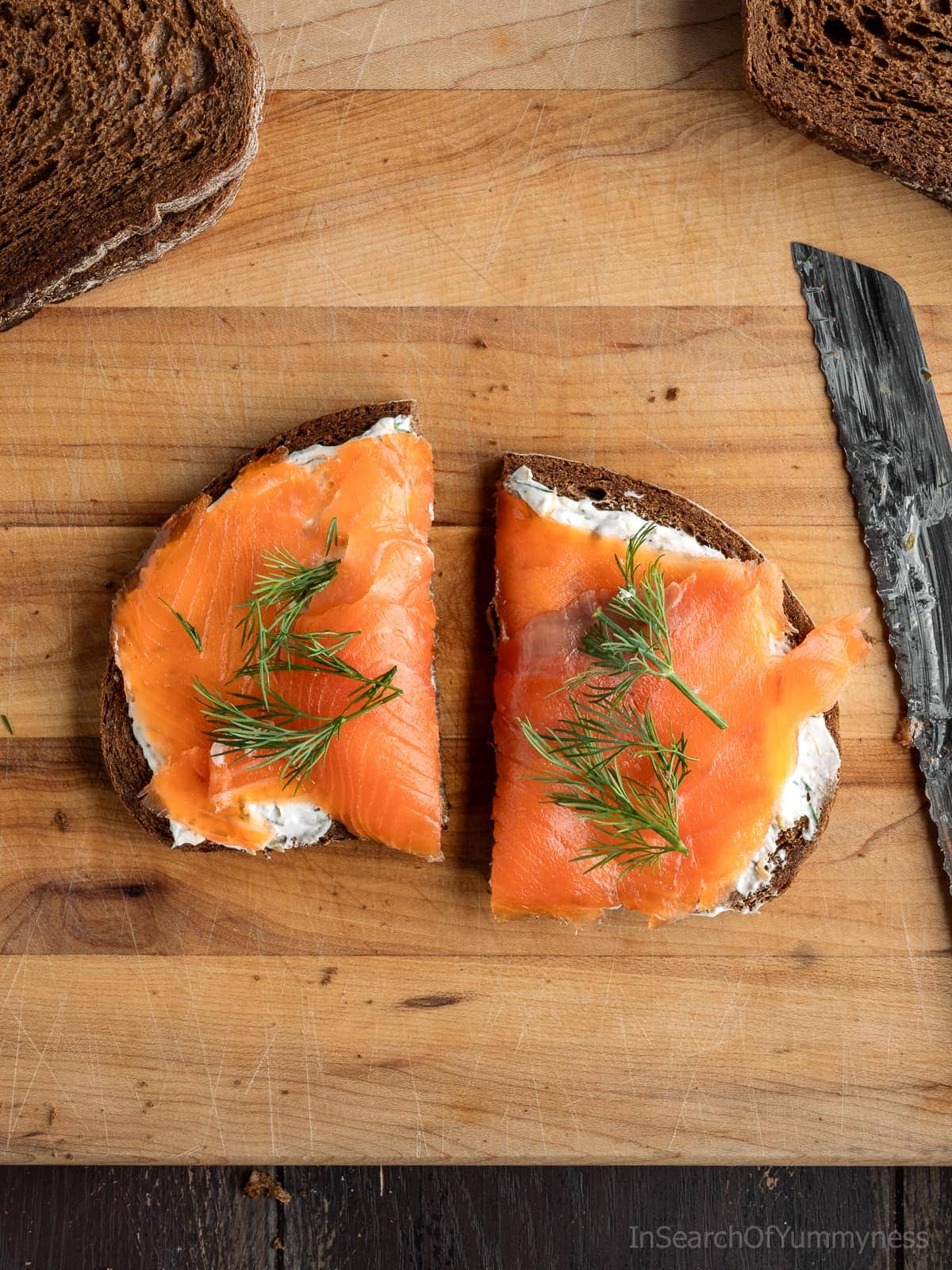 An open-faced smoked salmon tea sandwich, cut in half and topped with fresh dill, with a bread knife beside it.
