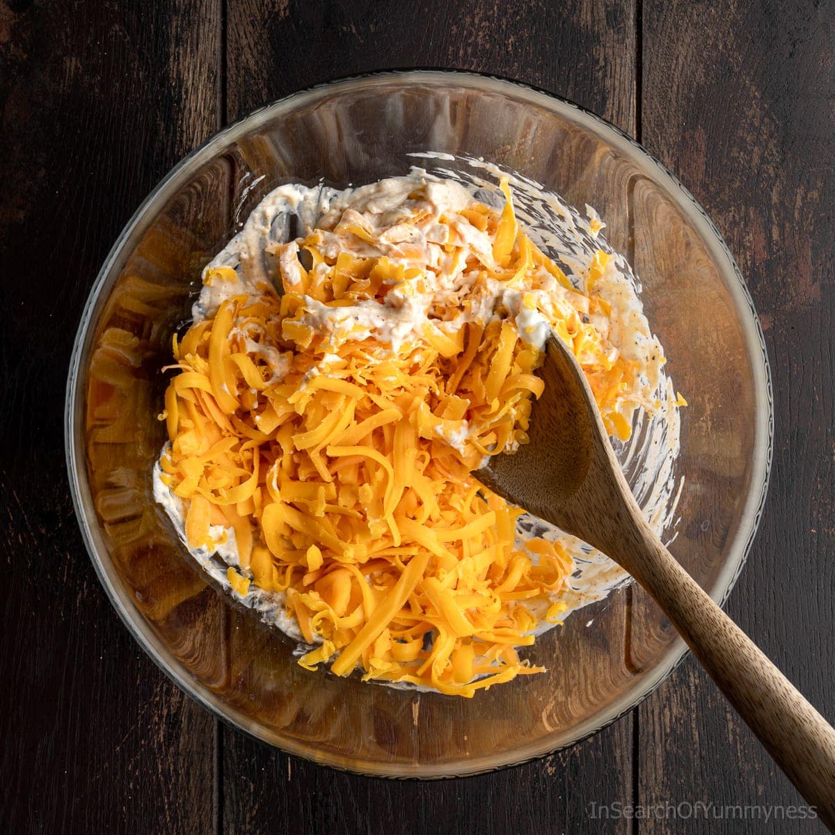 Adding shredded cheddar cheese to a clear mixing bowl containing a mayo, and cream cheese mixture, with a wooden spoon resting inside the bowl.