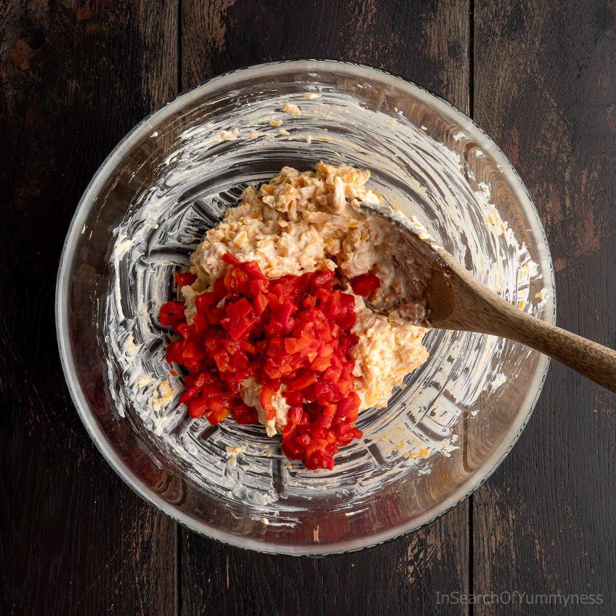 Adding pimentos to a clear mixing bowl containing a cheddar, mayo, and cream cheese mixture, with a wooden spoon resting inside the bowl.