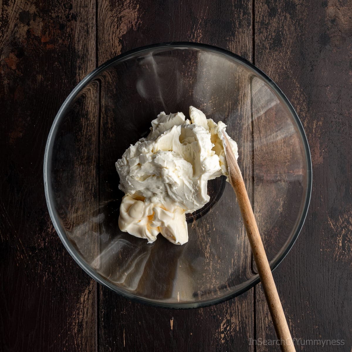 Cream cheese and mayonnaise together in a clear glass bowl with a wooden spoon.