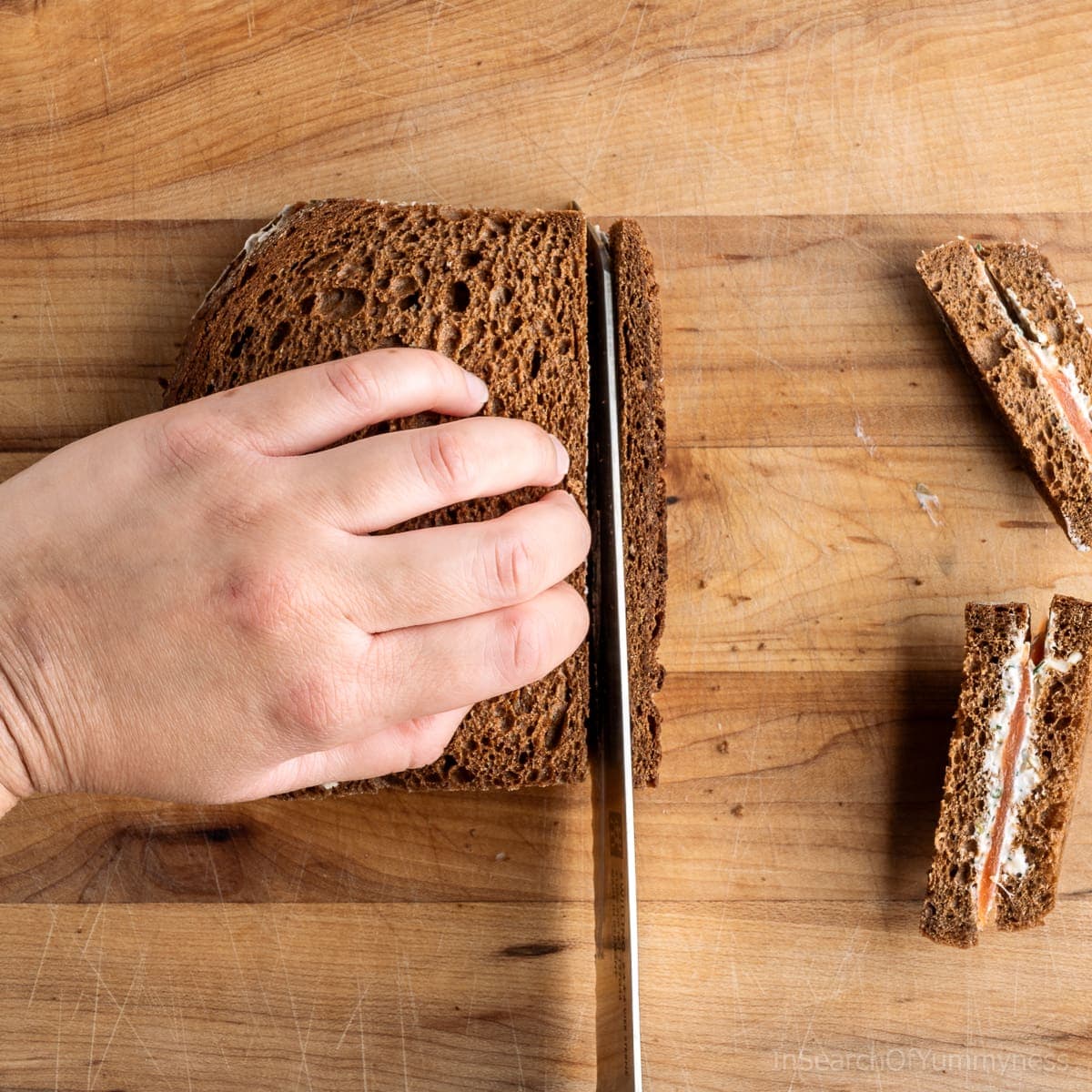 Hands using a bread knife to trim the crusts off a smoked salmon tea sandwich.