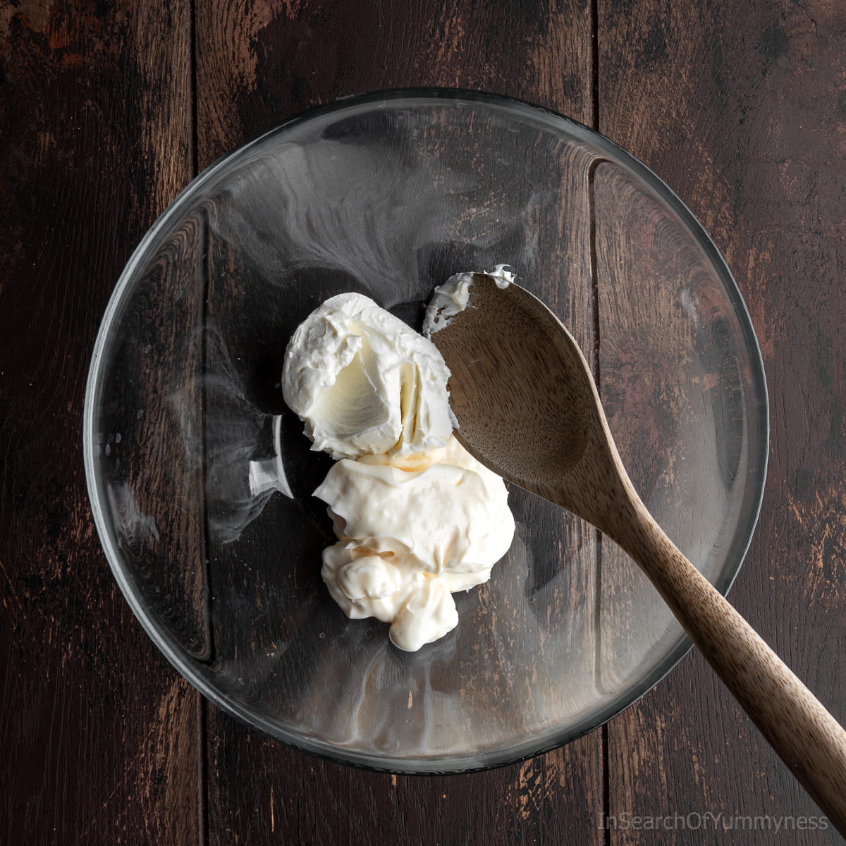 A clear mixing bowl filled with cream cheese and mayonnaise, with a wooden spoon resting inside.