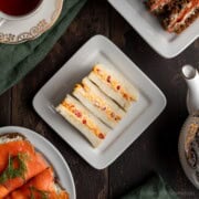Three pimento cheese tea sandwiches on a white plate next to a teapot and a cup of tea.