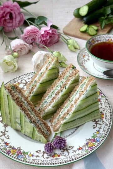 Four cucumber and herb tea sandwiches on an elegant plate next to a cup of tea and some roses.