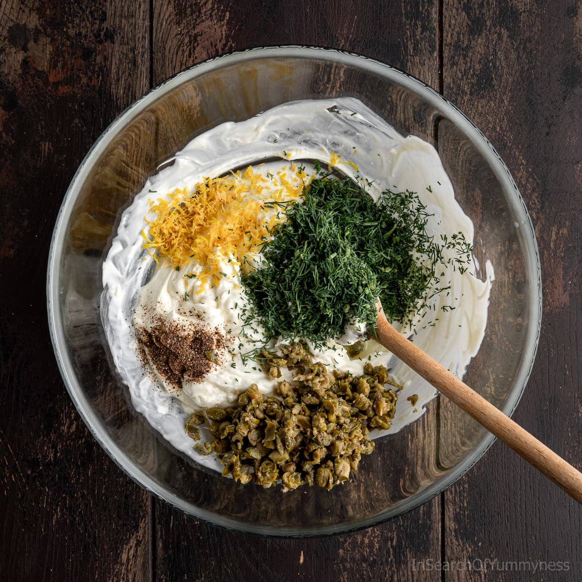 A clear glass bowl filled with a cream cheese and mayonnaise mixture, topped with fresh dill, lemon zest, capers, and black pepper, with a wooden spoon resting inside.