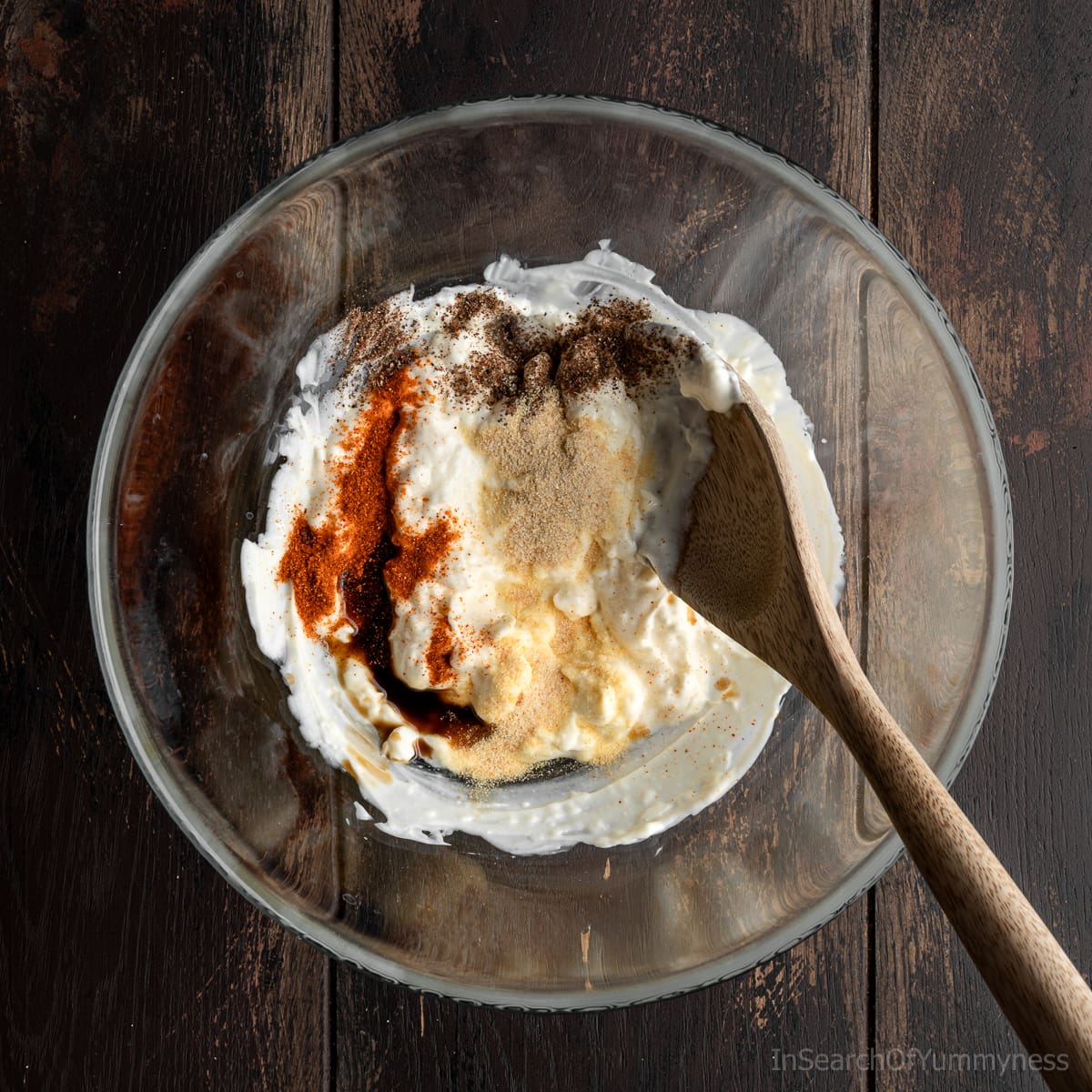 Adding seasonings to a clear mixing bowl containing a mayo and cream cheese mixture, with a wooden spoon resting inside the bowl.