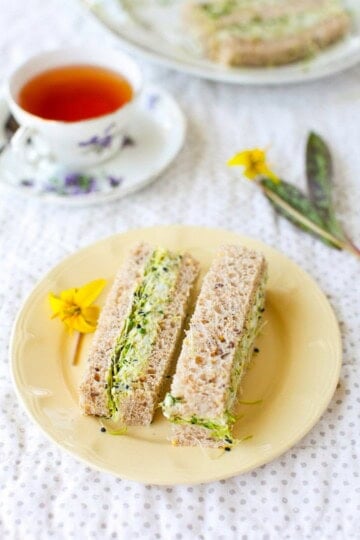 Two herbed avocado egg salad tea sandwiches on a yellow plate next to a cup of tea.