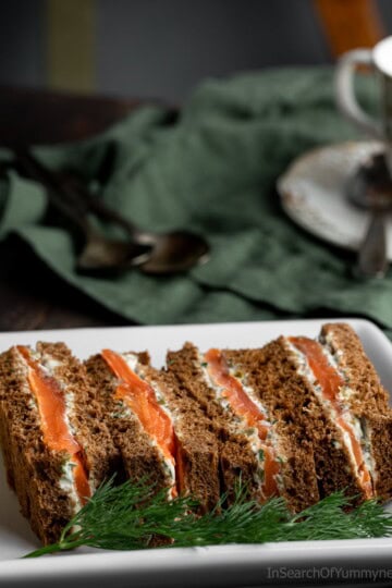 Four smoked salmon sandwiches on a white plate next to a cup of tea.
