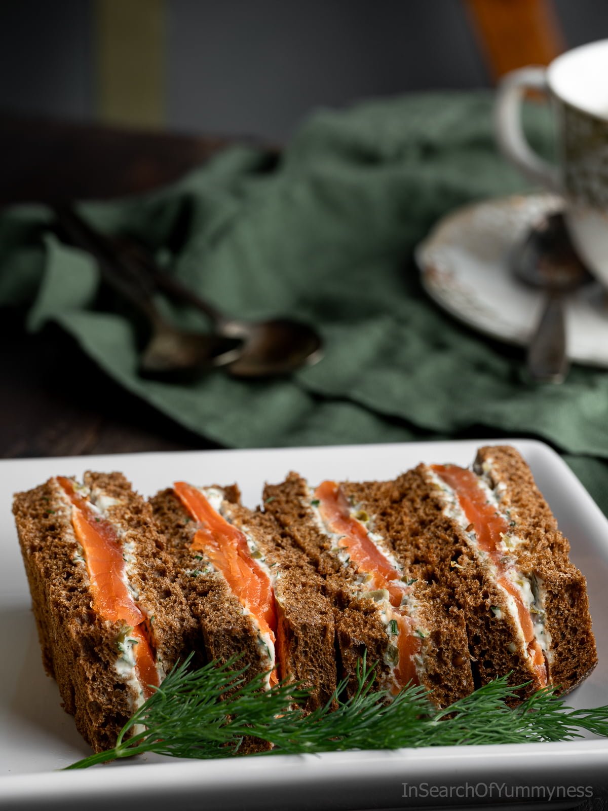 Four smoked salmon sandwiches on a white plate next to a cup of tea.