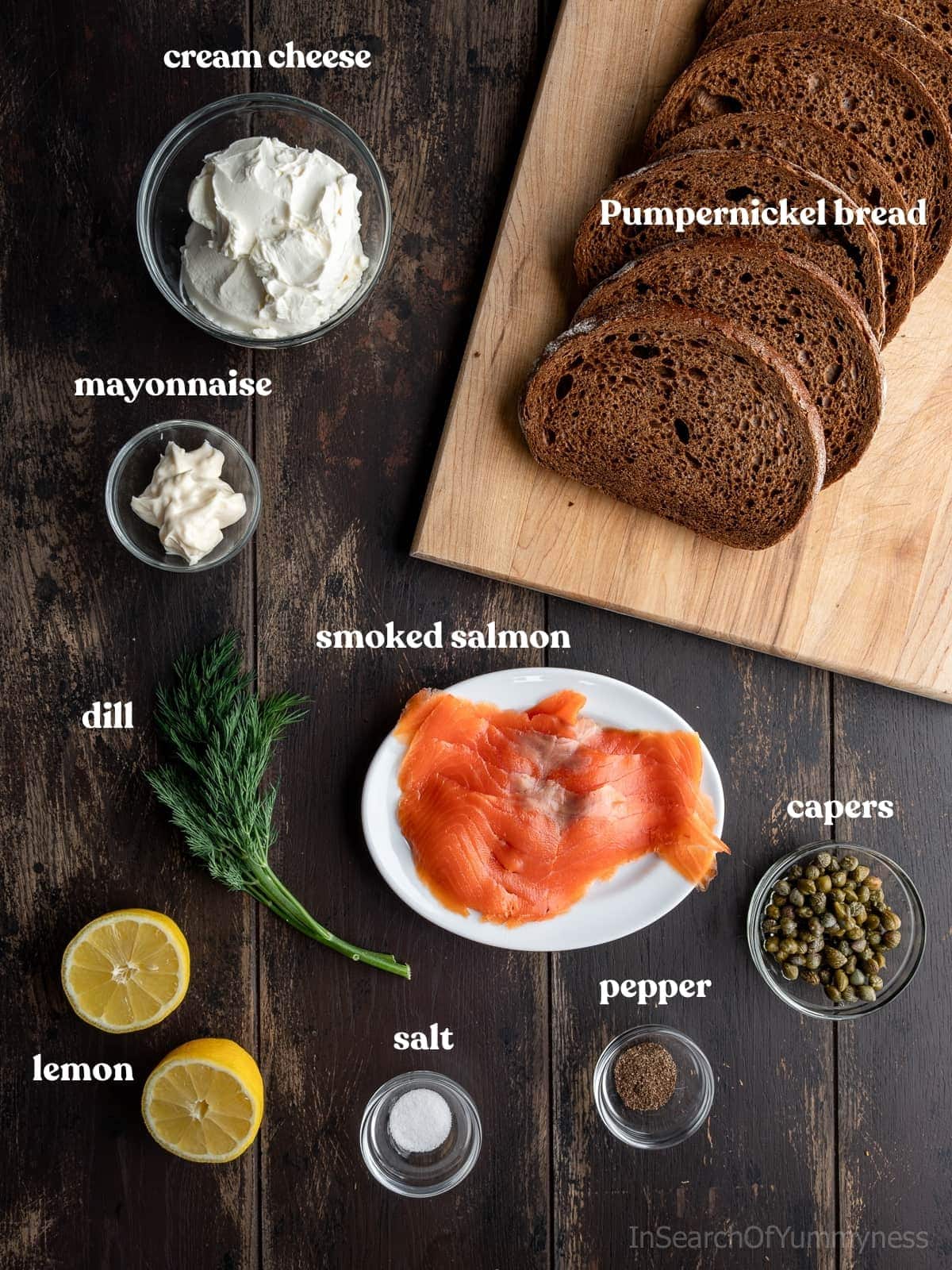 Ingredients used to make smoked salmon tea sandwiches layed out on a table. Ingredients include smoked salmon, Pumpernickel bread, cream cheese, capers and more.