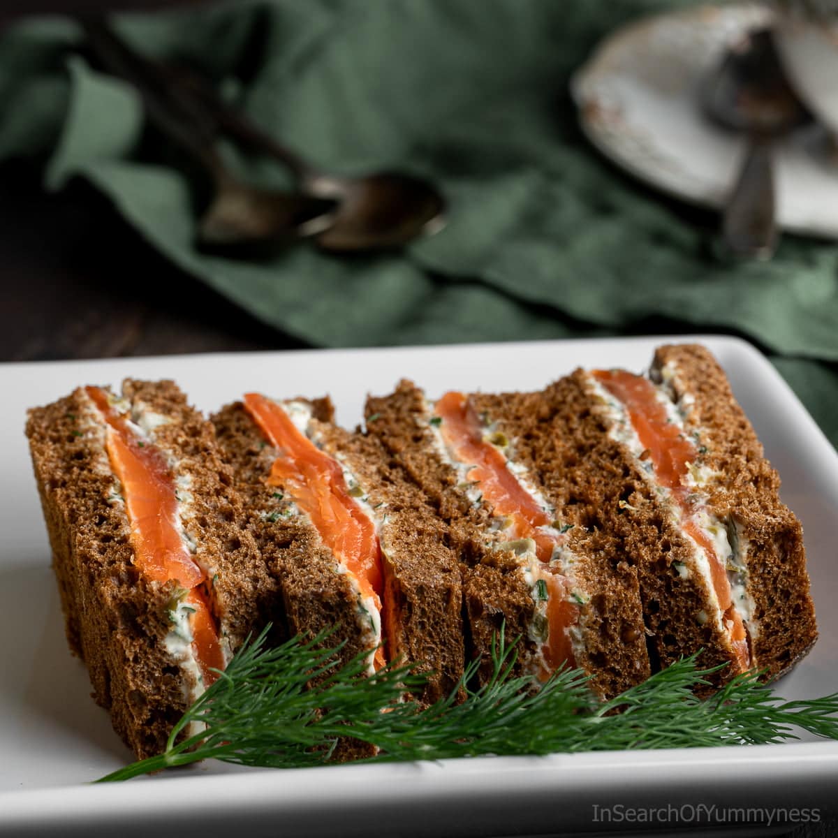 Four smoked salmon tea sandwiches on a white plate garnished with a sprig of dill.