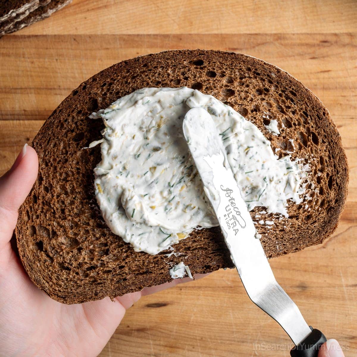 Spreading lemon dill cream cheese mixture onto a piece of Pumpernickel bread with an offset spatula.