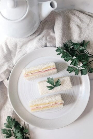 Three dainty ham and cheese tea sandwiches on a white plate next to a white pot of tea.