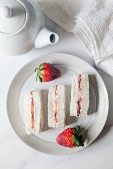 A white plate with three strawberry tea sandwiches next to two whole strawberries.