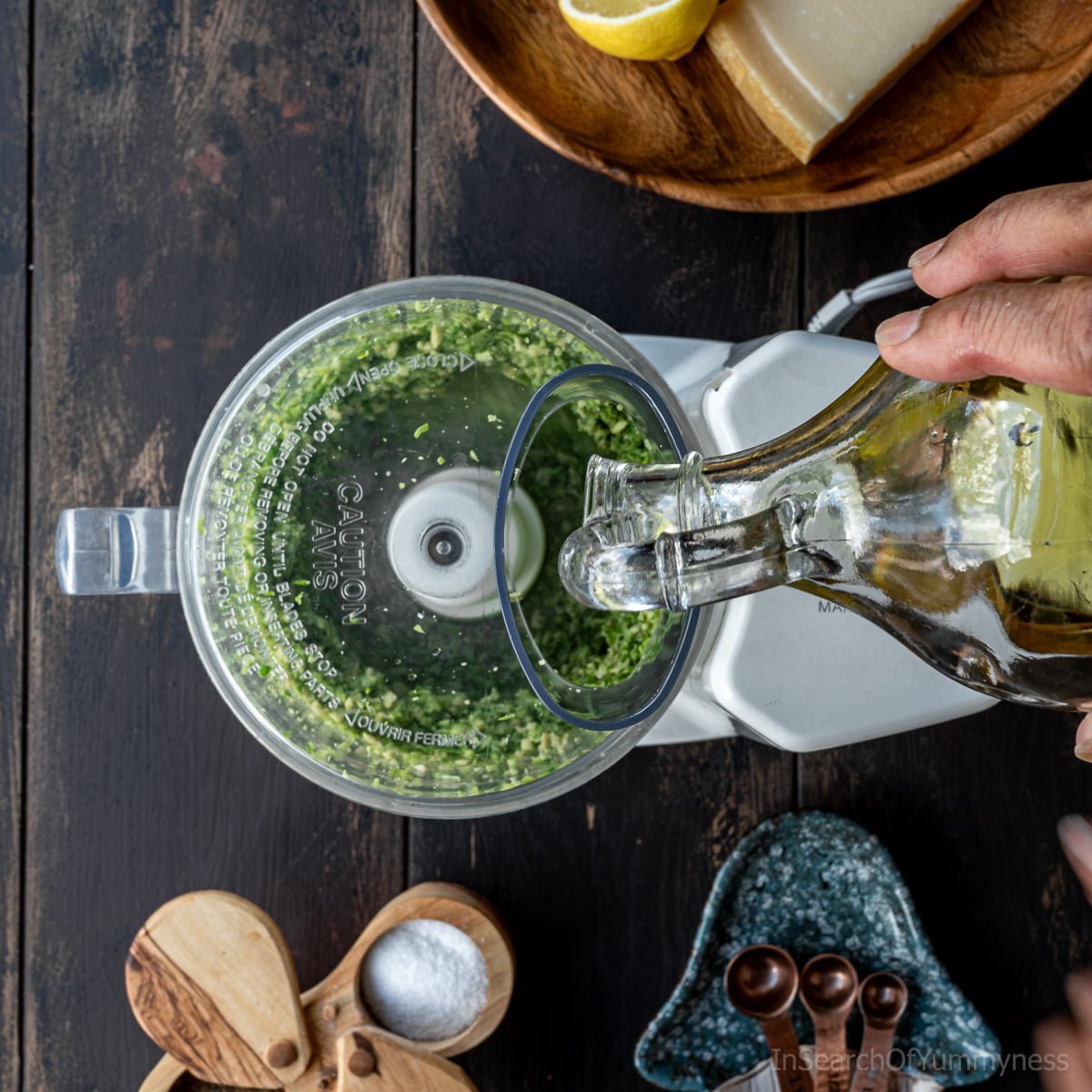 Pouring olive oil into a food processor that has garlic scapes and pumpkin seeds in it.