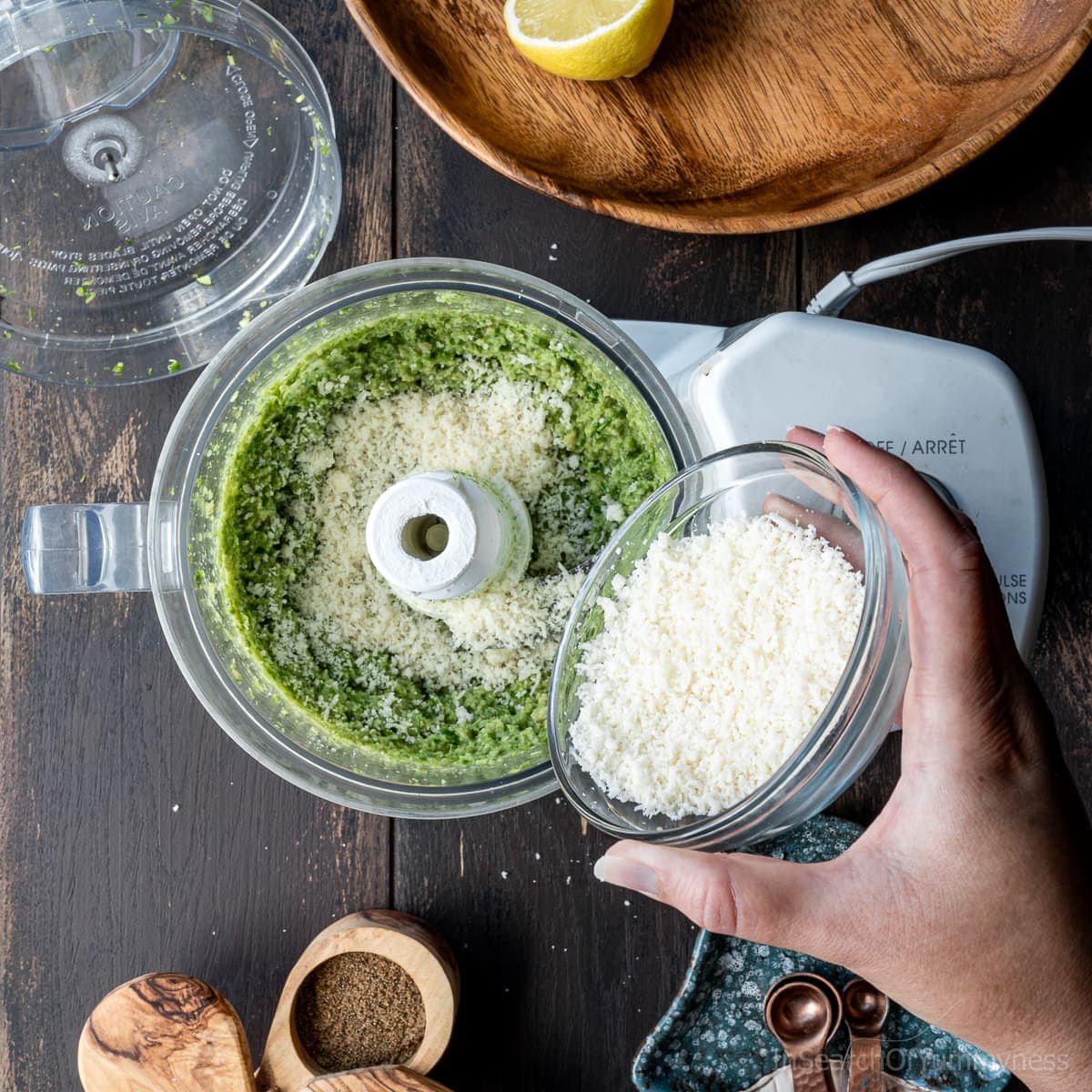 Adding a bowl of Parmesan cheese to garlic scape pesto in a food processor.