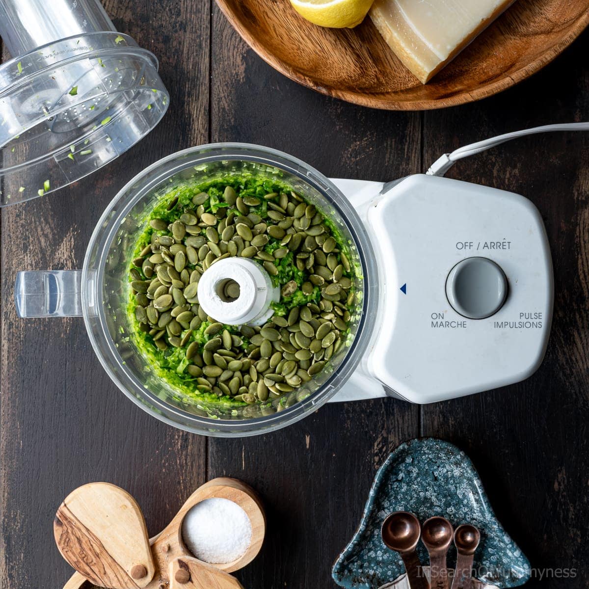 Pumpkin seeds sitting on top of chopped up garlic scapes inside of a food processor.