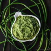 A garlic-shaped bowl full of garlic scape pesto, surrounded by fresh curly garlic scapes.
