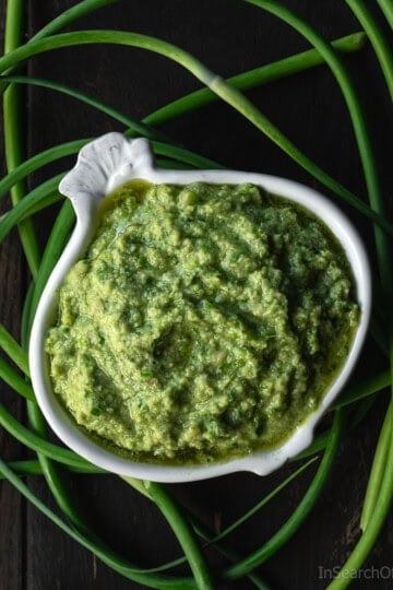 A garlic-shaped bowl full of garlic scape pesto, surrounded by fresh curly garlic scapes.