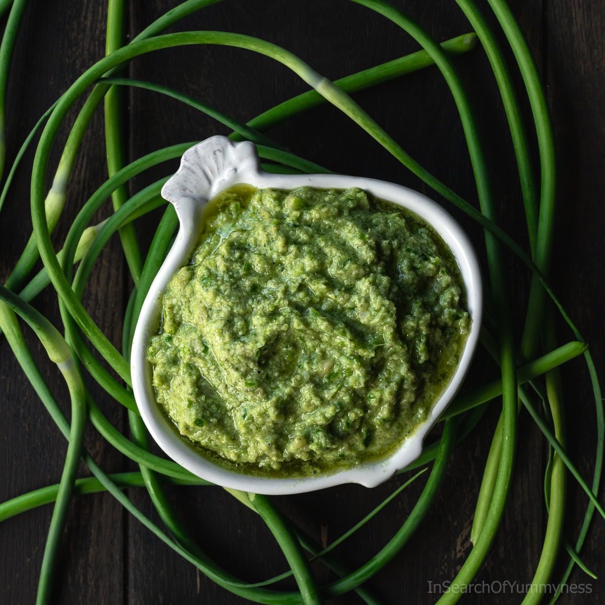 A garlic-shaped bowl full of garlic scape pesto, surrounded by fresh curly garlic scapes.