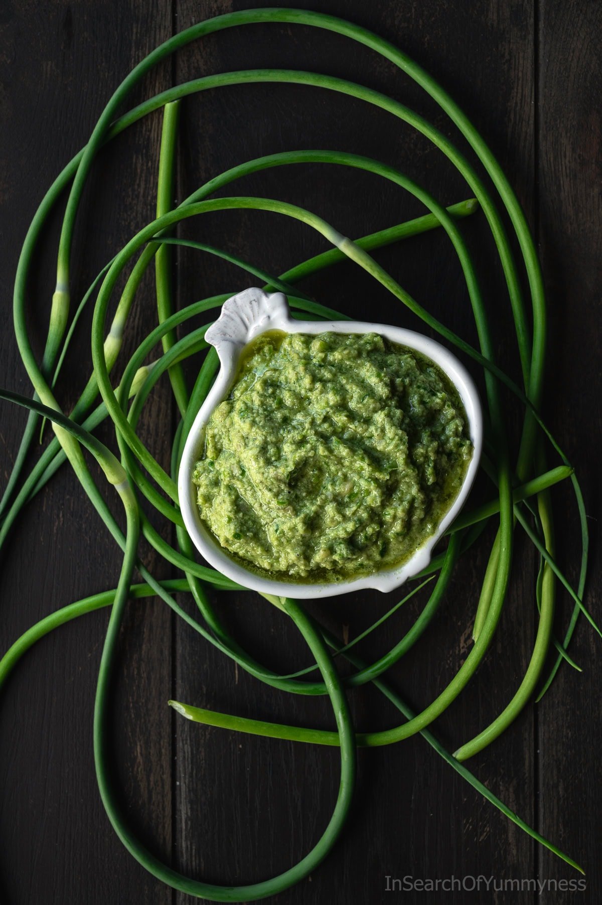 A garlic-shaped bowl full of garlic scape pesto, surrounded by fresh curly garlic scapes.