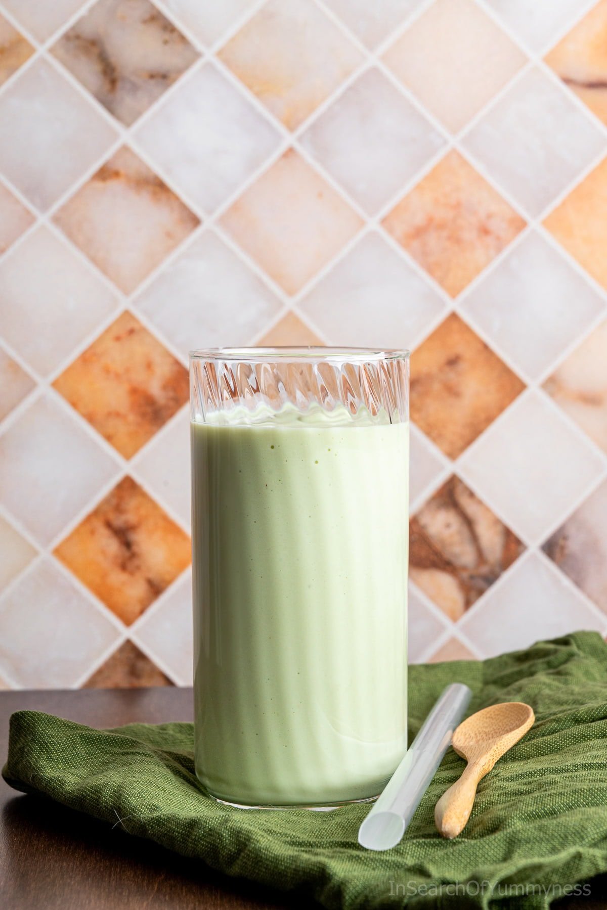 A tall glass of creamy Vietnamese avocado smoothie on a green napkin next to a thick straw and a wooden spoon.
