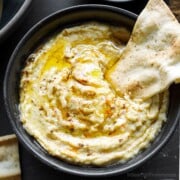 Creamy hummus, with a homemade pita chip dipped into it, in a black bowl.