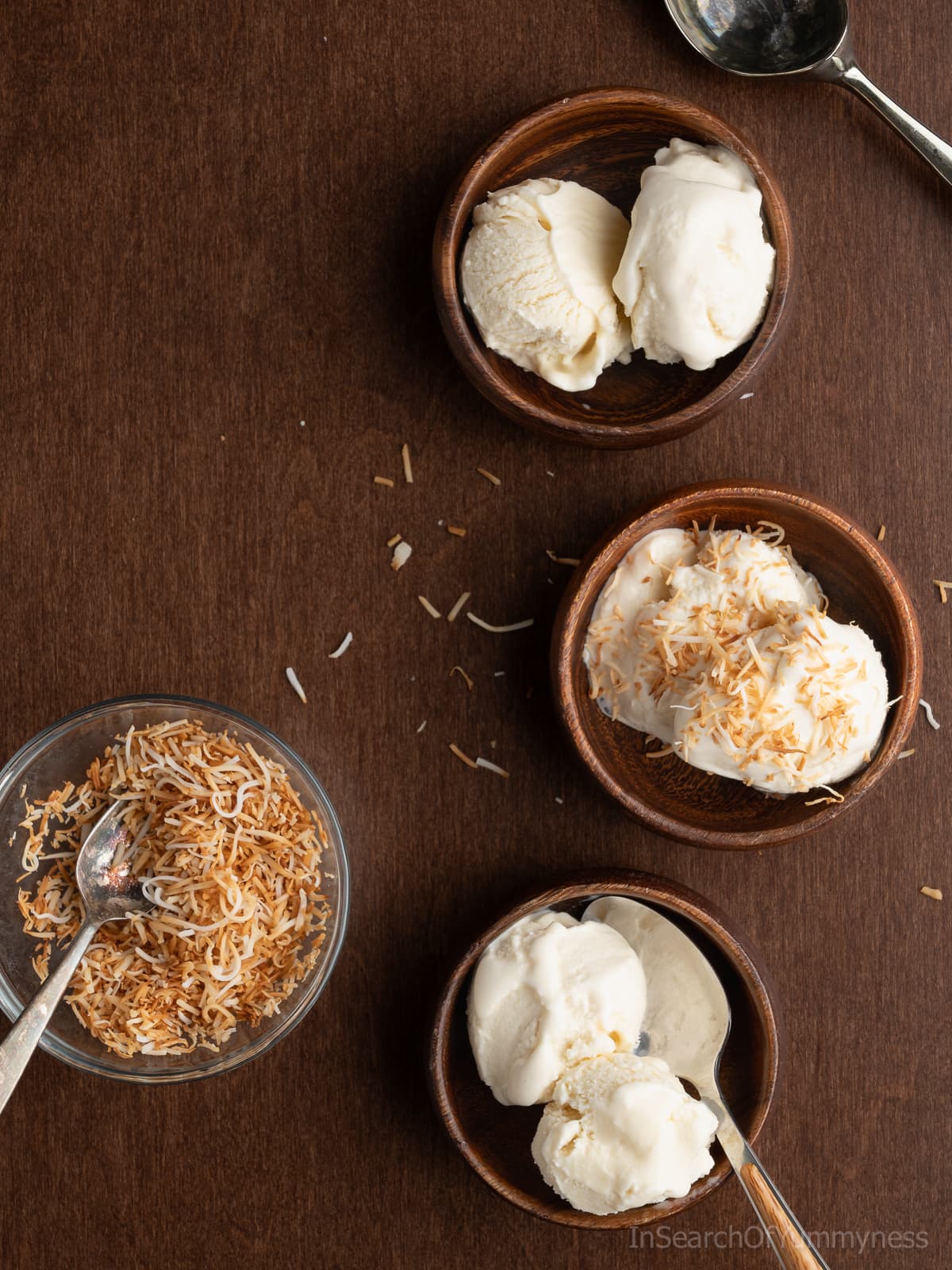 Three wooden bowls of Trini coconut ice cream next to a bowl of toasted coconut and an ice cream scoop.