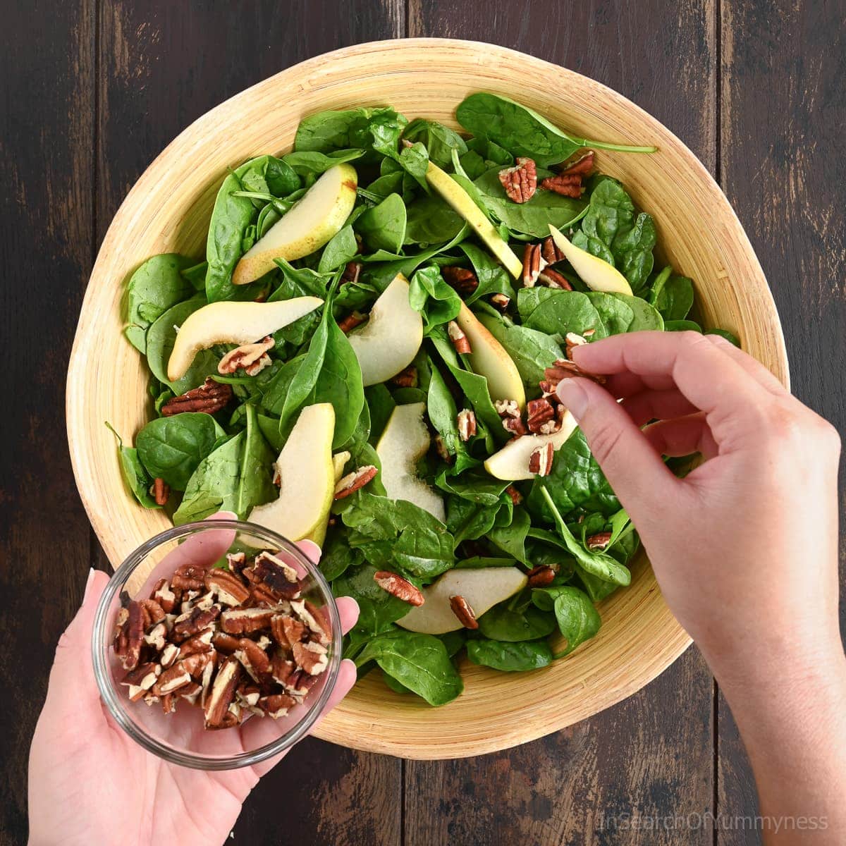 A person sprinkling chopped pecans over a wooden bowl filled with fresh spinach pear salad.