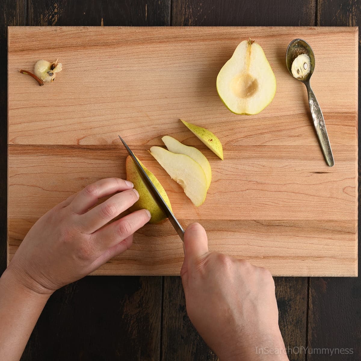 A pair of hands slicing a ripe pear into thin wedges on a wooden cutting board, with the cored fruit, spoon, and seeds nearby.
