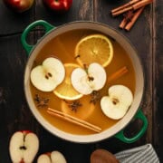A green Le Creuset Dutch oven filled with apple cider, warm spices, orange slices and apple slices.