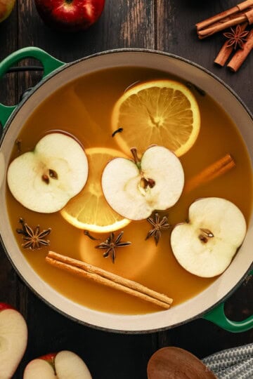 A green Le Creuset Dutch oven filled with apple cider, warm spices, orange slices and apple slices.