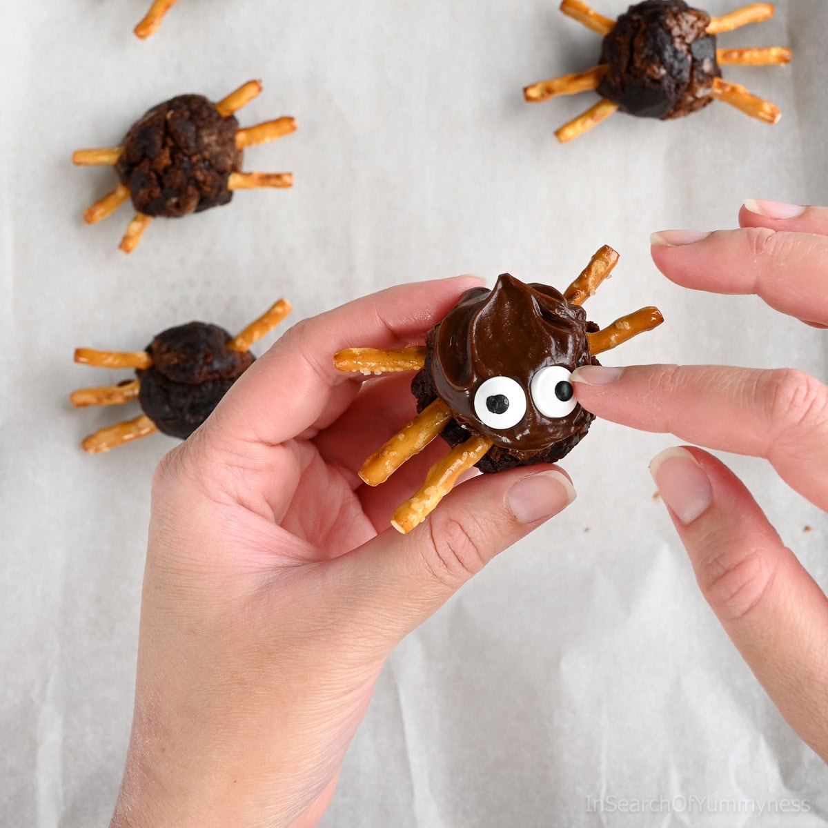 Hands decorating a spider brownie by pressing candy eyeball sprinkles into chocolate frosting on a round brownie ball with pretzel stick legs, with more finished spider brownies in the background on parchment paper.