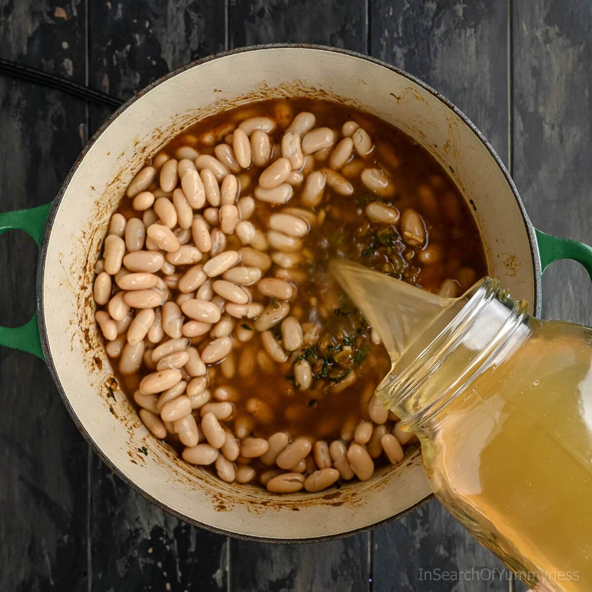 Pouring chicken stock from a glass jar into a Dutch oven full of onions, spices and white kidney beans.