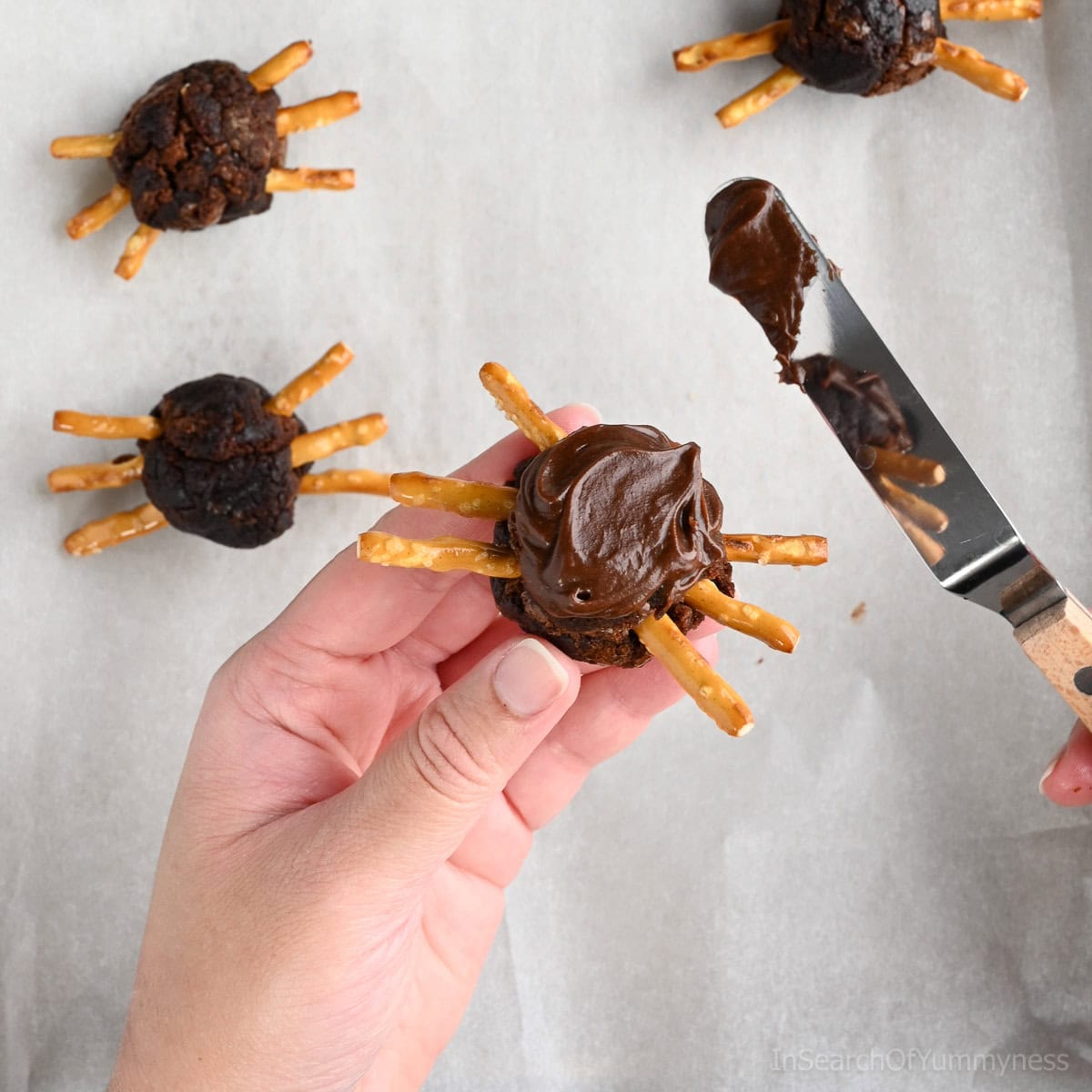 Hands decorating a spider brownie by applying chocolate frosting on a round brownie ball with pretzel stick legs, with more finished spider brownies in the background on parchment paper.