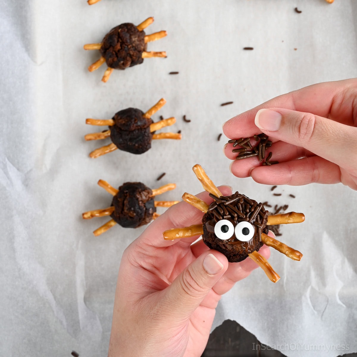 Hands decorating a spider brownie by adding chocolate sprinkles onto chocolate frosting on a round brownie ball with pretzel stick legs, with more finished spider brownies in the background on parchment paper.