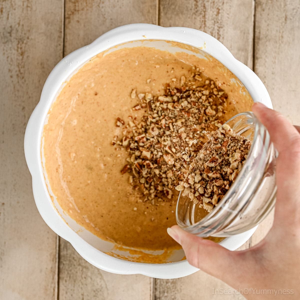 Adding finely chopped toasted pecans from a glass bowl into a white pumpkin shaped bowl that has pumpkin waffle batter in it.