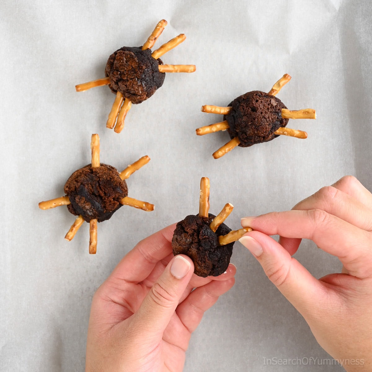 Hands decorating a spider brownie by inserting pretzel stick pieces into a round brownie ball to make spider legs. There are three other spider brownies in progress on parchment paper.