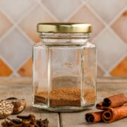 A small glass jar with a gold lid containing an allspice substitute spice mix. The jar sits on a wooden surface with whole nutmeg, cloves, and cinnamon sticks around it.