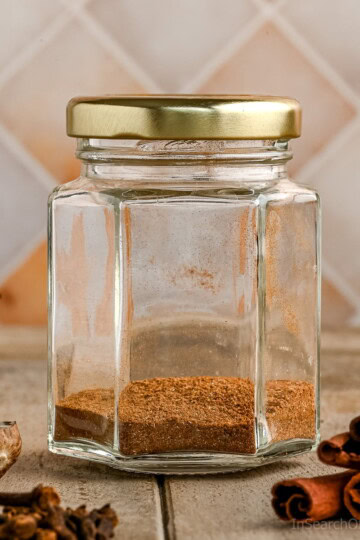 A small glass jar with a gold lid containing an allspice substitute spice mix. The jar sits on a wooden surface with whole nutmeg, cloves, and cinnamon sticks around it.