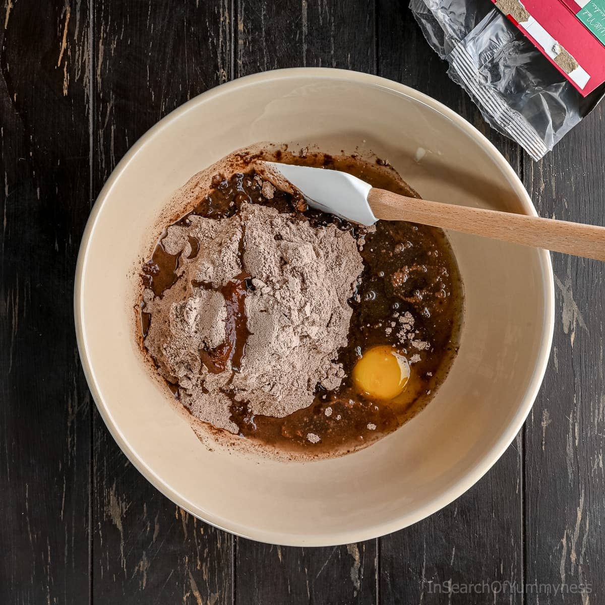 The contents of a box of brownie mix in a peach-coloured bowl with oil, water and an egg. Waiting to be combined with a spatula sitting in the bowl.