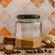 A jar of a homemade pumpkin spice blend with a black lid, next to various whole spices including cinnamon, clove, nutmeg, allspice and a knob of fresh ginger.