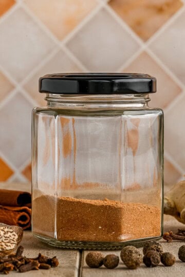 A jar of a homemade pumpkin spice blend with a black lid, next to various whole spices including cinnamon, clove, nutmeg, allspice and a knob of fresh ginger.