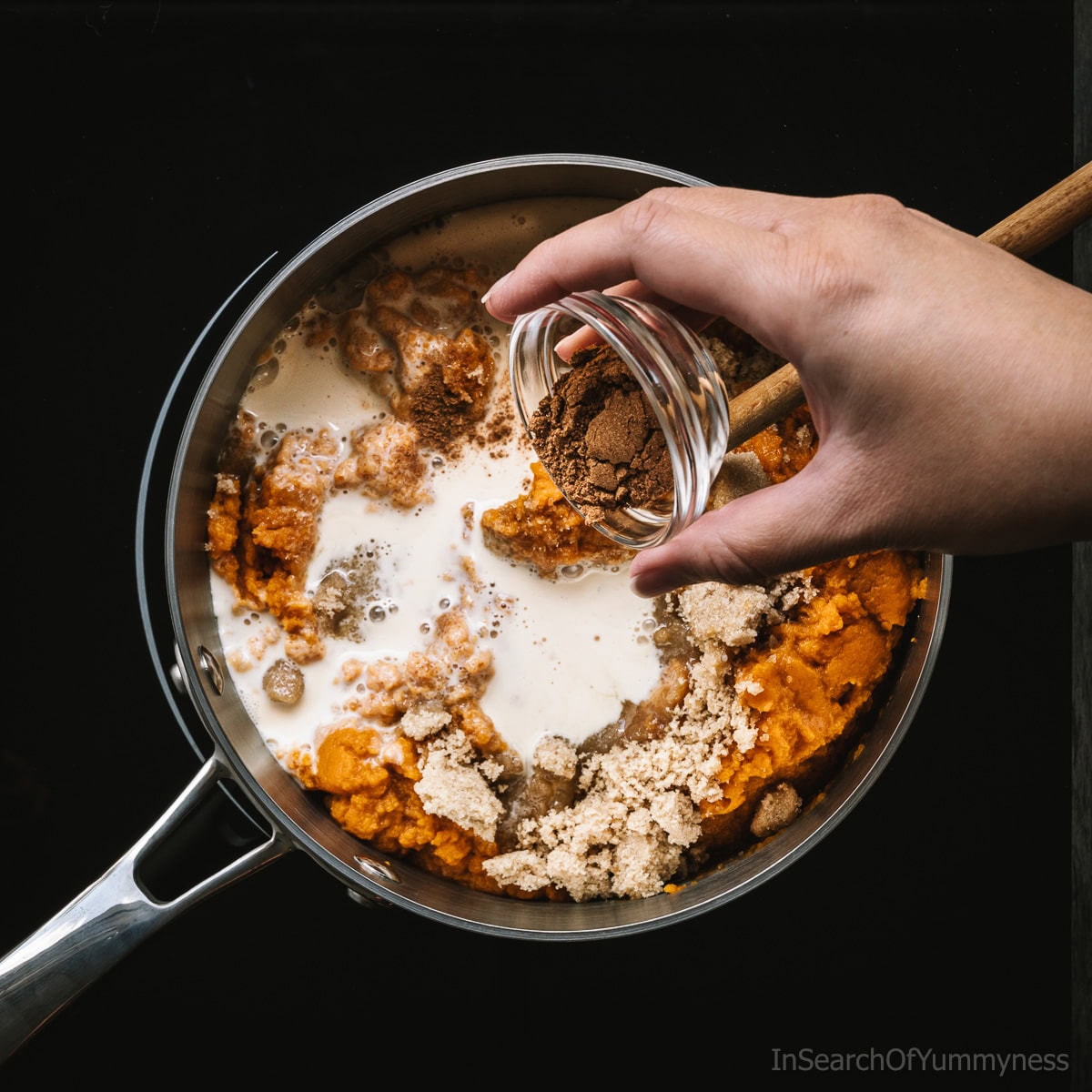 A steel saucepan full of ingredients to make pumpkin pie filling for crepes, including pumpkin puree and brown sugar. A hand is tipping a clear bowl of spices into the pot.