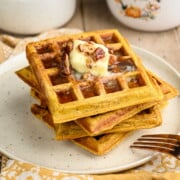 A stack of 4 golden pecan pumpkin waffles on a beige plate, topped with a pat of butter and drizzled with maple pecan syrup.