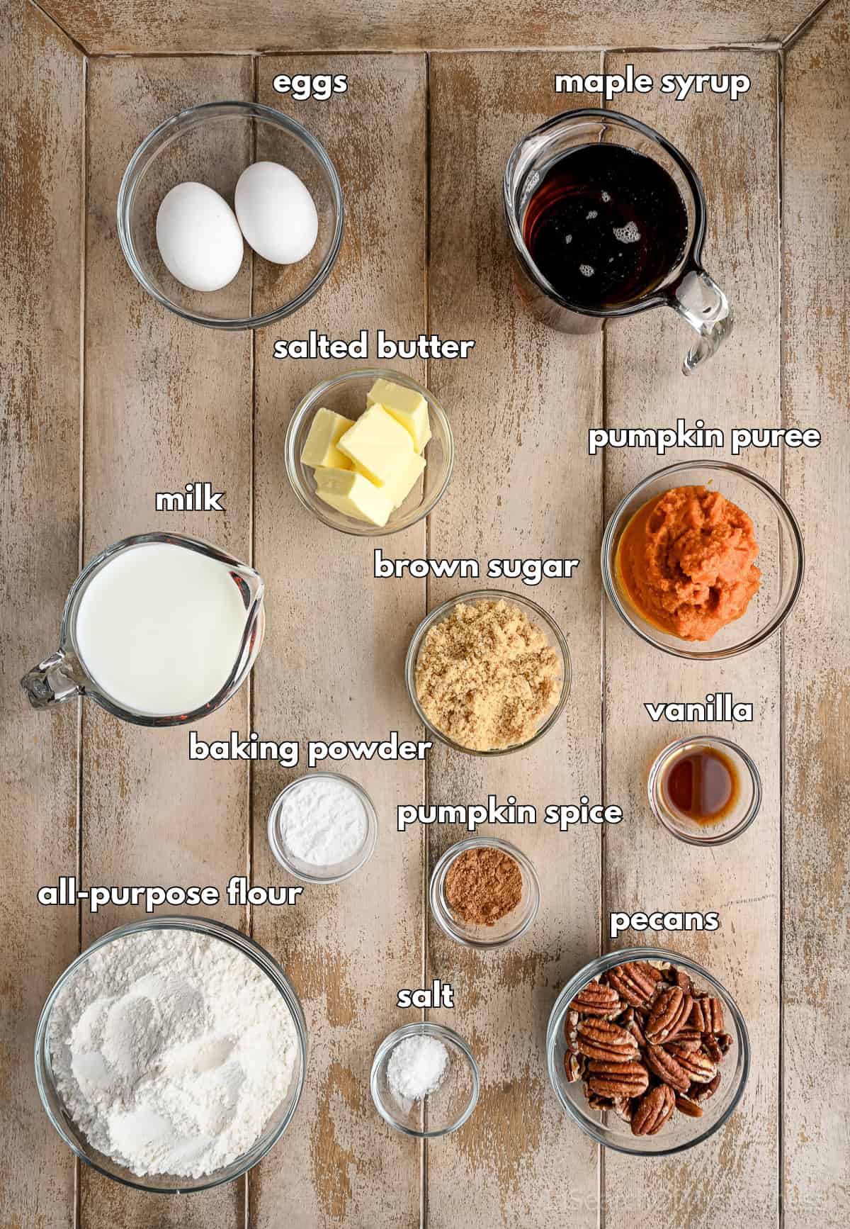 Alt text: Ingredients for pumpkin pecan waffles arranged on a wooden surface, including flour, milk, eggs, butter, brown sugar, pumpkin puree, maple syrup, pecans, vanilla, pumpkin spice, baking powder, and salt.