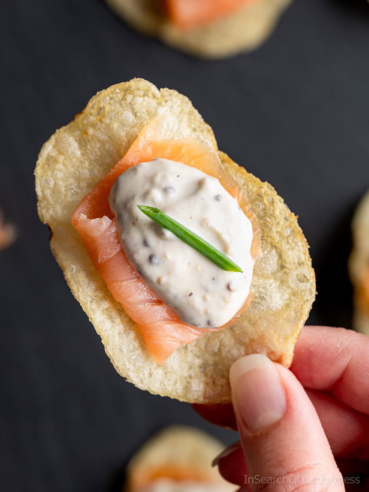 A hand holding a potato chip canap&eacute; topped with a slice of smoked salmon, a dollop of creamy sauce, and a small piece of chive on top.